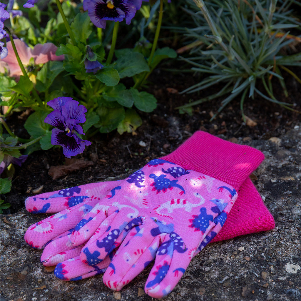 Kent and Stowe gardening gloves for children in pink and blue with dinosaurs resting by flowerbed